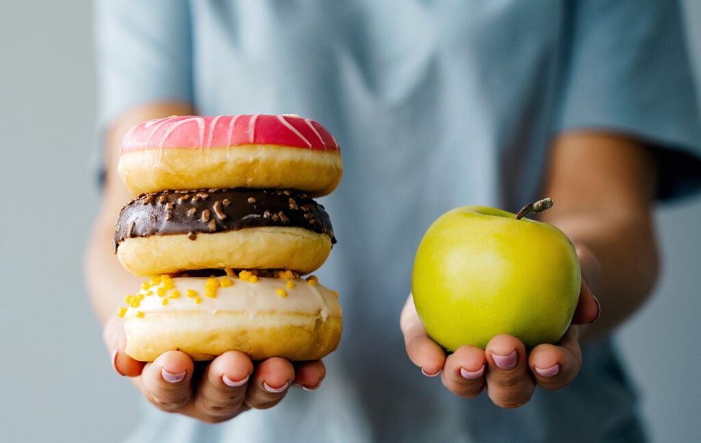 Hands holding donuts and a fresh apple, illustrating food choices in a plant-forward eating pattern that supports vascular and arterial health.