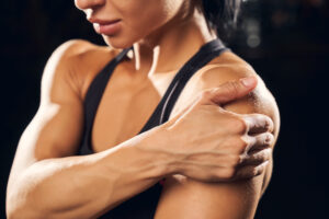 Muscular woman in gym attire holding her shoulder, close-up on arm and shoulder suggesting sore muscle. Mitozz, (-)-epicatechin